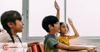 Three children sitting at desks. Two raising their hands