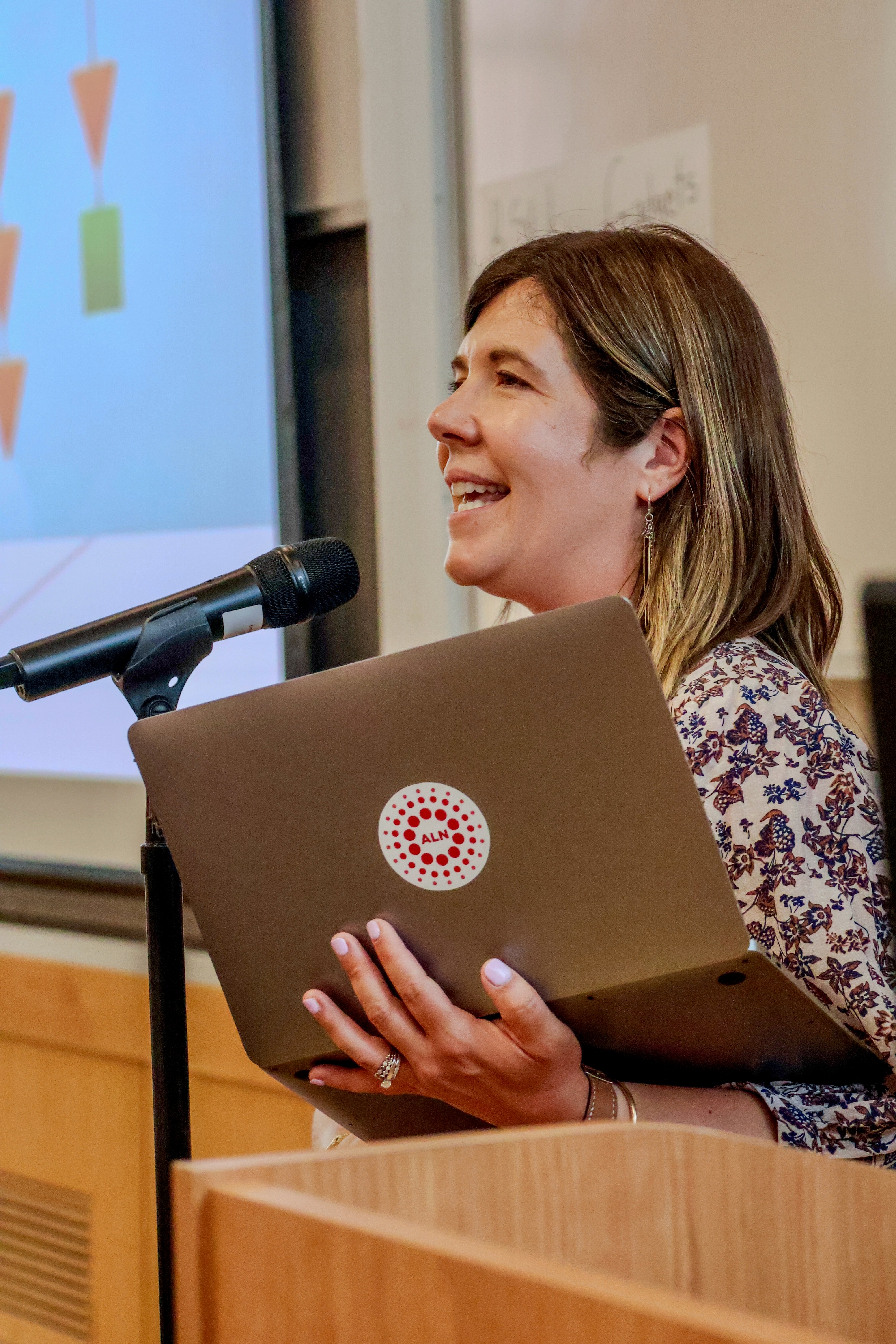 Ashley Marlow speaking in front of an audience. Ashley is speaking into a microphone and holding a laptop.