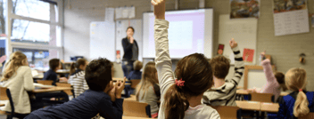 A young girl raises her hand in the classroom while her teacher looks on.