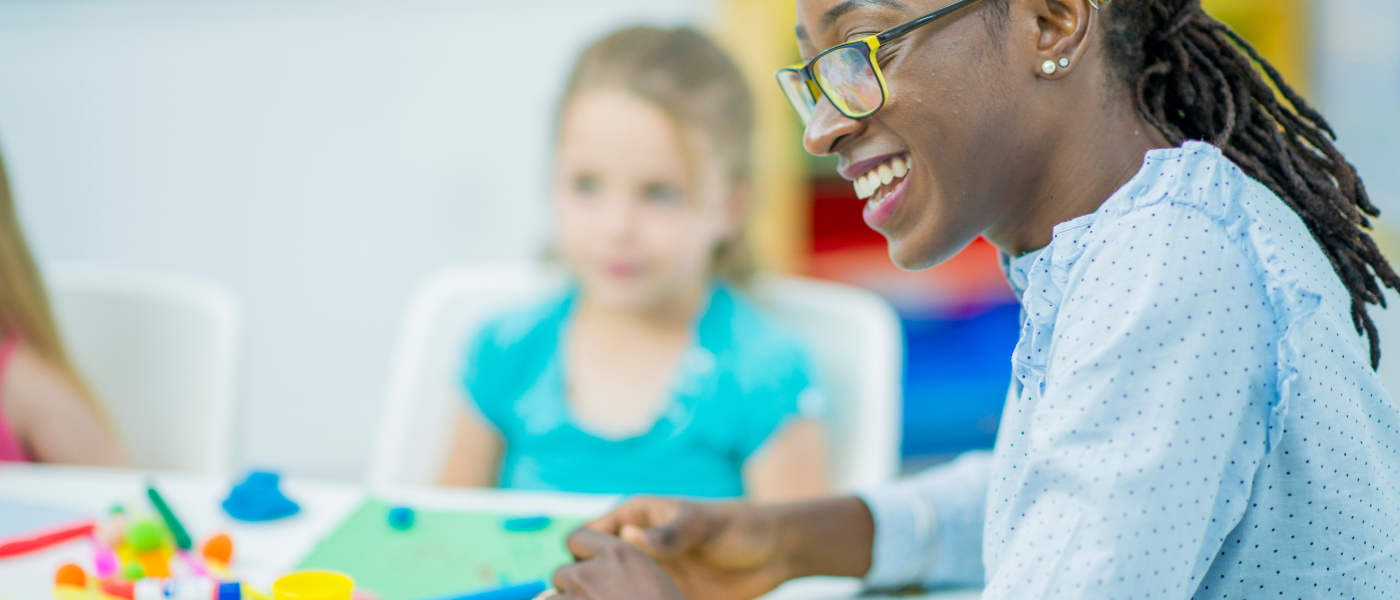 A teacher smiles at the class while a student looks on.
