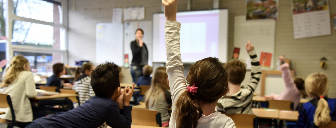 A young girl raises her hand in the classroom while her teacher looks on.